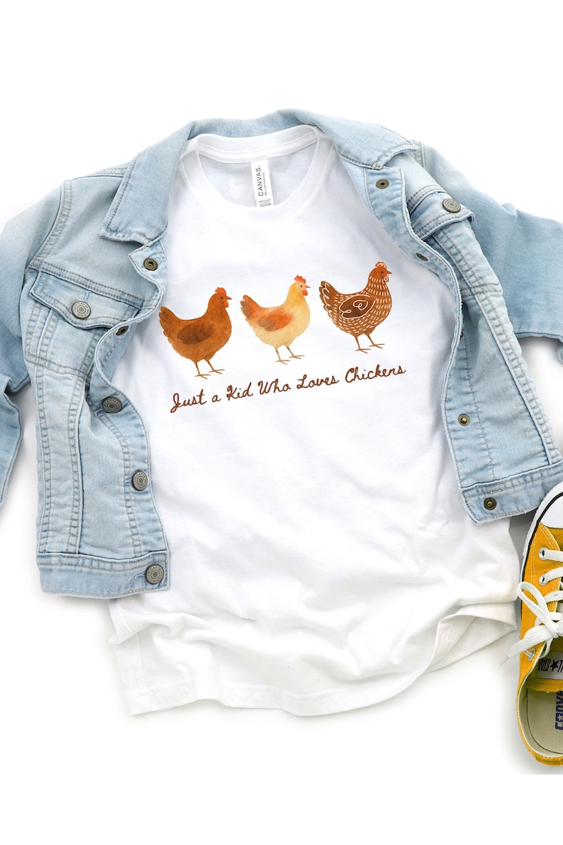 May include: White t-shirt with three watercolor chickens and the text "Just a Kid Who Loves Chickens." A light blue denim jacket is draped over the shirt. Yellow sneakers are visible in the bottom right corner.