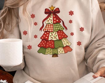 May include: Sand-colored sweatshirt featuring a Christmas tree design. The tree is composed of patchwork patterns in red, green, and white, topped with a gold star and red bow. Red snowflakes are scattered around the tree.