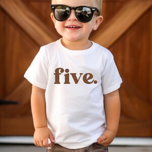 May include: White t-shirt with the word "five" in brown lettering. The child is wearing black sunglasses and brown pants. The background is a wooden door.