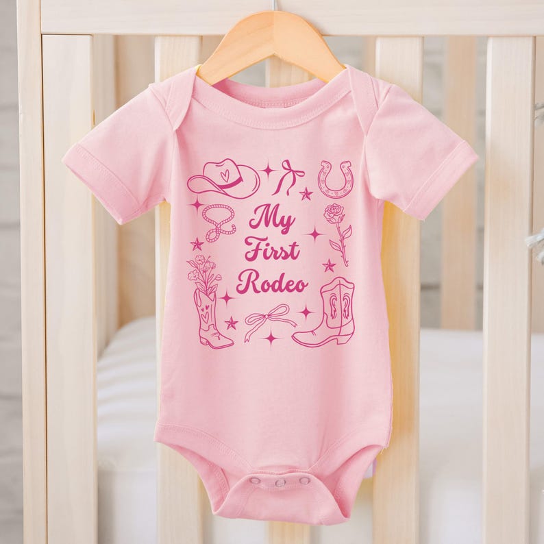 May include: Pink baby onesie with a pink graphic design that says "My First Rodeo" and includes a cowboy hat, horseshoe, boots, and other western-themed imagery.