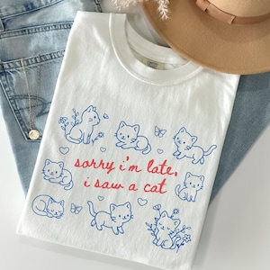 May include: White t-shirt with a crew neck featuring blue cat illustrations and the text "sorry i'm late, i saw a cat" in red. The shirt is folded on top of blue jeans, with a gold necklace and a tan hat.