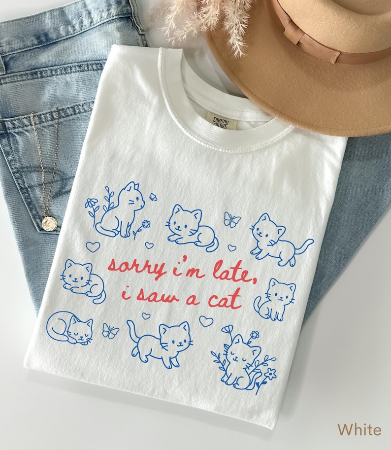 May include: White t-shirt with a crew neck featuring blue cat illustrations and the text "sorry i'm late, i saw a cat" in red. The shirt is folded on top of blue jeans, with a gold necklace and a tan hat.