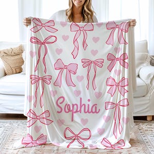 May include: A white blanket featuring a pattern of pink bows and hearts. The name "Sophia" is written in pink script. The blanket is held up by a person.