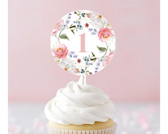May include: A white cupcake with white frosting topped with a round cake topper featuring a floral design in shades of pink, white, and blue. The cake topper has the number "1" in pink.