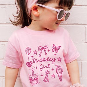 May include: Pink t-shirt with a pink graphic design that says "Birthday Girl" and includes a heart, cupcake, ice cream cone, butterfly, star, and party hat.