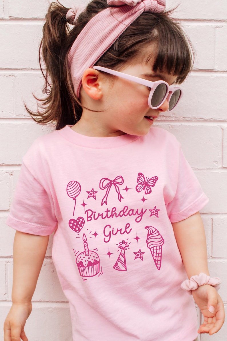May include: Pink t-shirt with a pink graphic design that says "Birthday Girl" and includes a heart, cupcake, ice cream cone, butterfly, star, and party hat.