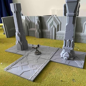 May include: A grey, 3D printed miniature set for tabletop gaming. The set includes two tall pillars with intricate designs and a square tile with a patterned surface. A miniature figure stands on the tile.
