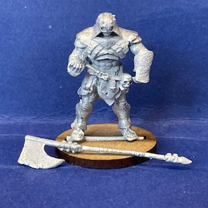 Puede incluir: Una figura en miniatura de plata de un guerrero que lleva un casco y una armadura, sosteniendo un arma. La figura está de pie sobre una base de madera. Un hacha de plata está tirada en el suelo junto a la base.