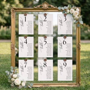 May include: A gold-coloured framed seating plan featuring nine white cards, each with a table number and guest names. The cards are secured with brown circular fasteners. Floral decorations are placed on the frame.