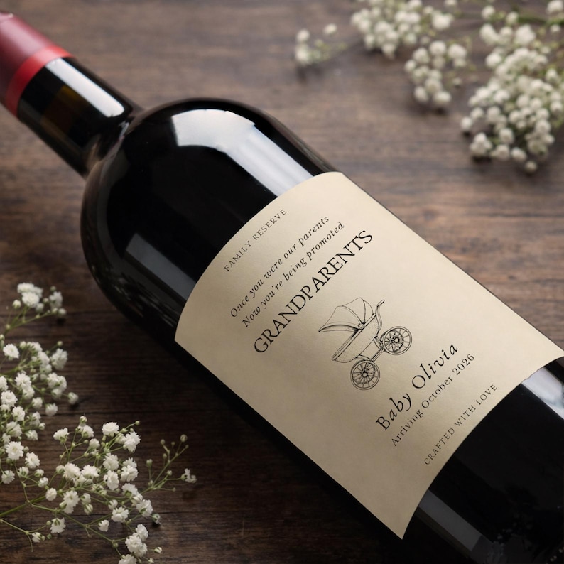 Pu&ograve; includere: Una bottiglia di vino rosso scuro con un'etichetta personalizzata. L'etichetta recita "GRANDPARENTS" con un'illustrazione di un passeggino e "Baby Olivia Arriving October 2026". La bottiglia &egrave; su una superficie di legno con fiori bianchi.