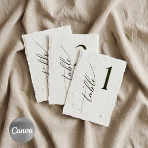 May include: Three handmade white paper table number cards with deckled edges. Each card has the word "table" in elegant script and a number in a bold font. The cards are arranged on a beige linen surface.