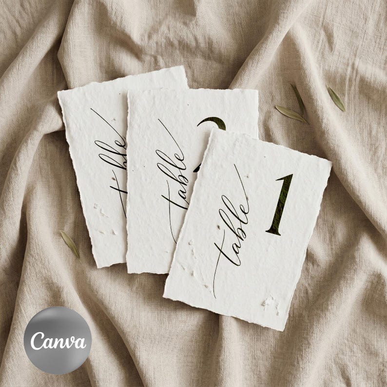 May include: Three handmade white paper table number cards with deckled edges. Each card has the word "table" in elegant script and a number in a bold font. The cards are arranged on a beige linen surface.