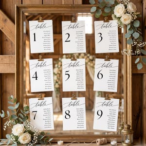 Pode incluir: Um espelho com moldura de madeira exibe nove cartões brancos com números de mesa e nomes de convidados, presos com clipes dourados. Os cartões são dispostos em uma grade, com detalhes florais e uma estética rústica, ideal para um casamento.