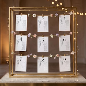 May include: A gold metal frame displaying nine cards with guest names, each numbered and labeled "table." The cards are clipped to a chain adorned with small flowers. Fairy lights are strung around the frame, creating a warm, inviting ambiance.