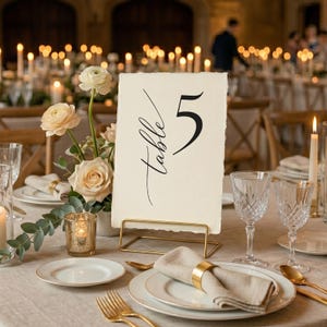 May include: Elegant table setting with a cream-colored table number card that reads "Table 5" in black script. The table is set with gold-rimmed plates, gold cutlery, and crystal glassware. A floral arrangement and candles add to the ambiance.