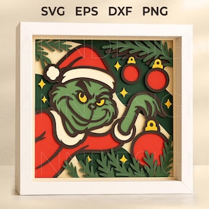 May include: A framed, layered paper art piece depicting a green character in a Santa hat, reaching for a red bauble. The artwork has a Christmas theme, with additional red baubles, yellow stars, and green foliage. The top of the frame displays the text "SVG EPS DXF PNG".