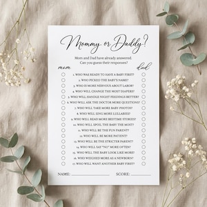 May include: A white rectangular baby shower game card titled Mommy or Daddy? with black text. The card features seventeen numbered questions about parenting, with circular checkboxes for mom and dad, and lines for name and score at the bottom.