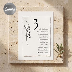 May include: A white table number card with the number 3 in black, and the word "table" in cursive. The card lists names and is set on a textured stone surface. A sprig of greenery is in the lower right corner.