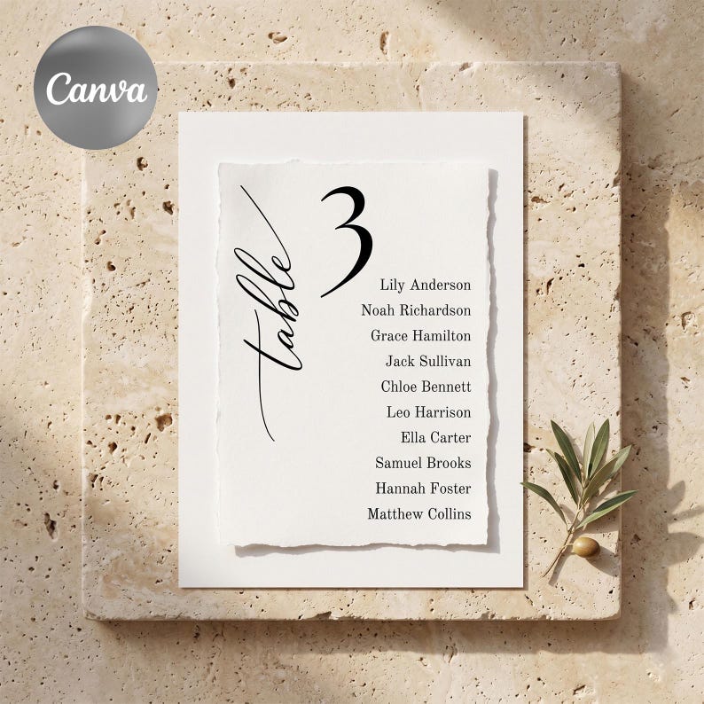 May include: A white table number card with the number 3 in black, and the word "table" in cursive. The card lists names and is set on a textured stone surface. A sprig of greenery is in the lower right corner.