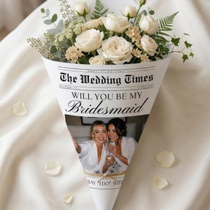 Puede incluir: Un ramo de rosas blancas y vegetación envuelto en un cono estilo periódico. El texto dice "The Wedding Times" y "Will You Be My Bridesmaid". Se ve una foto de dos mujeres.