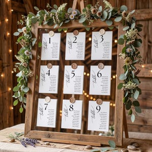 May include: A wooden framed seating chart with nine numbered cards, each listing names and sealed with a wax stamp. The frame is decorated with greenery and flowers, perfect for a wedding or event.