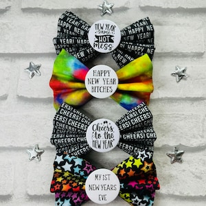 May include: Three colorful bow ties with different New Year's themed buttons. The top bow tie is black with white text, the middle bow tie is tie-dye, and the bottom bow tie is black with colorful stars. The buttons say "New Year Same Hot Mess", "Happy New Year Bitches", "Cheers to the New Year", and "My 1st New Years Eve".