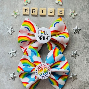 May include: Two rainbow fabric hair bows with white clouds and rainbow patterns. One bow has a white button with the text "Full of Pride" and a heart. The other bow has a white button with the text "All for Love Love for All" and a peace sign.