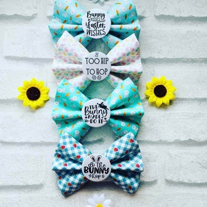 May include: Four Easter-themed hair bows with different patterns and sayings. The bows are made of blue and white gingham, white with pastel coloured polka dots, and blue with white bunnies and carrots. The sayings on the bows are "Bunny Kisses Easter Wishes", "Too Hip To Hop", "The Bunny Made Do It", and "Do The Bunny Hop".