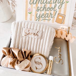 May include: A gift box containing a beige ribbed pouch with the name Sarah, a gold-toned compact mirror with the letter S, a gold-toned lip balm, two satin scrunchies, and a gray water bottle with the name Sarah and a peach claw clip.