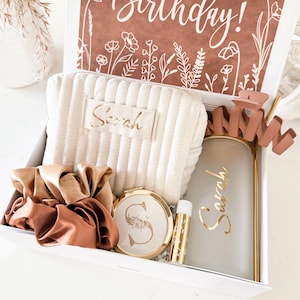 May include: A white gift box filled with beauty items. Includes a beige quilted cosmetic bag, gold-toned compact mirror, lip balm, scrunchies, and a frosted tumbler with a straw. A card with "Happy Birthday!" is visible.