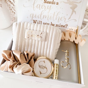 May include: A gift box with a princess theme, containing a beige cosmetic bag, a gold compact mirror, a lip balm, a tumbler, and a scrunchie. The box lid has text that reads "Every Princess Needs a Fairy Godmother. Will you be mine?"