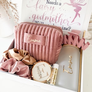 May include: A gift box with a pink theme, containing a pink velvet cosmetic bag with "Emma" in gold lettering, a compact mirror, lip balm, scrunchies, and a frosted glass tumbler. A card with text "Every Princess Needs a Fairy Godmother" is visible.