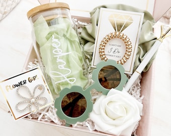 Puede incluir: Una caja de regalo rosa con un tarro con el nombre "Harper", gafas de sol verdes, un bolígrafo con una parte superior en forma de diamante, una goma para el pelo en espiral, un broche de flores y una rosa blanca. La caja está llena de papel blanco.
