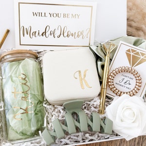 May include: A gift box filled with items for a maid of honor. The box includes a glass jar with a green lid, a white jewelry box with a gold letter K, a green hair clip, a gold pen, a gold hair tie, a white card with a gold diamond, a white card with the text "I can't say I Do without you!" and a white rose.