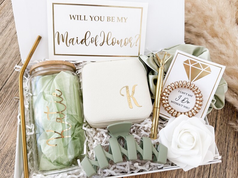 May include: A gift box filled with items for a maid of honor. The box includes a glass jar with a green lid, a white jewelry box with a gold letter K, a green hair clip, a gold pen, a gold hair tie, a white card with a gold diamond, a white card with the text "I can't say I Do without you!" and a white rose.
