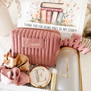 May include: A gift box with a pink theme, containing a corduroy cosmetic bag, scrunchies, a compact mirror, a lip balm, and a candle. The box lid features a floral illustration with books and the text "THANK YOU FOR BEING PART OF MY STORY!"