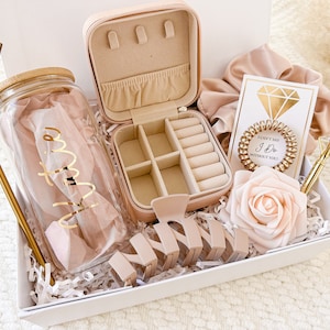 May include: A gift box filled with items for a bridal shower or wedding. The box includes a clear glass water bottle with a gold straw, a pink jewelry box, a pink hair scrunchie, a pink rose, a gold hair clip, and a card that says "I can't say I do without you".