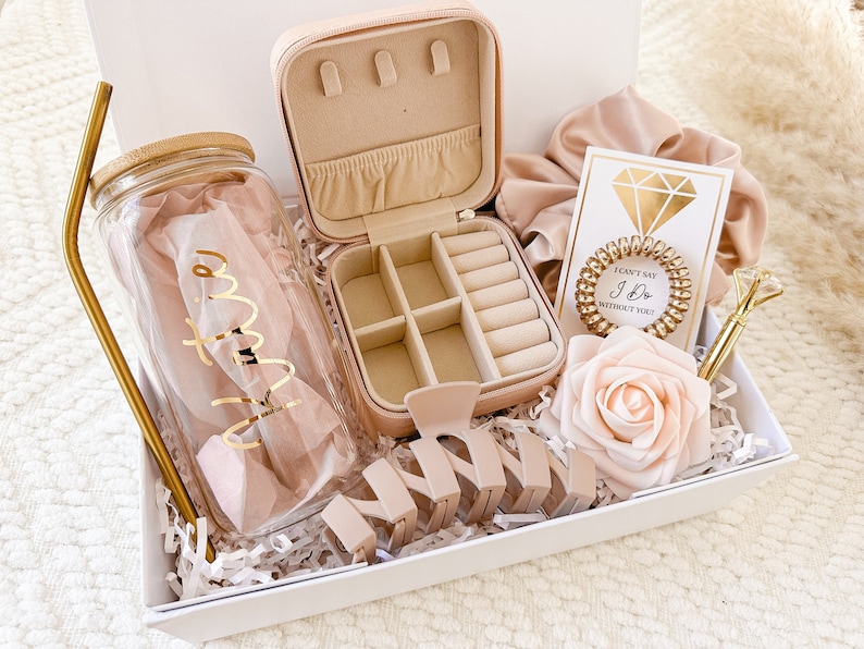 May include: A gift box filled with items for a bridal shower or wedding. The box includes a clear glass water bottle with a gold straw, a pink jewelry box, a pink hair scrunchie, a pink rose, a gold hair clip, and a card that says "I can't say I do without you".