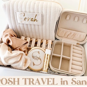 May include: A gift set in a white box. It includes a beige travel jewellery case, a matching cosmetic bag with "Sarah" in gold lettering, scrunchies, a compact mirror, and lip gloss. The text "POSH TRAVEL in Sand" is at the bottom.