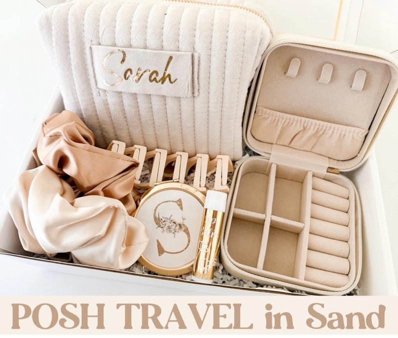 May include: A gift set in a white box. It includes a beige travel jewellery case, a matching cosmetic bag with "Sarah" in gold lettering, scrunchies, a compact mirror, and lip gloss. The text "POSH TRAVEL in Sand" is at the bottom.