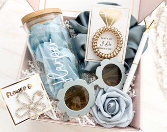 May include: A gift box filled with blue items, including a glass jar with the name "Harper", sunglasses, a hair tie, a pen, a flower pin, and a paper rose. The box is lined with white paper and blue tissue paper.