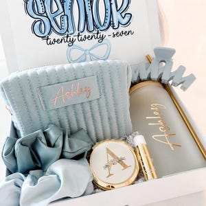 Peut inclure: Coffret cadeau blanc contenant une trousse de maquillage bleu clair, des chouchous, un miroir compact, un baume &agrave; l&egrave;vres et un gobelet. Le couvercle de la bo&icirc;te porte l'inscription "Senior" en bleu, avec le texte "twenty twenty - seven".