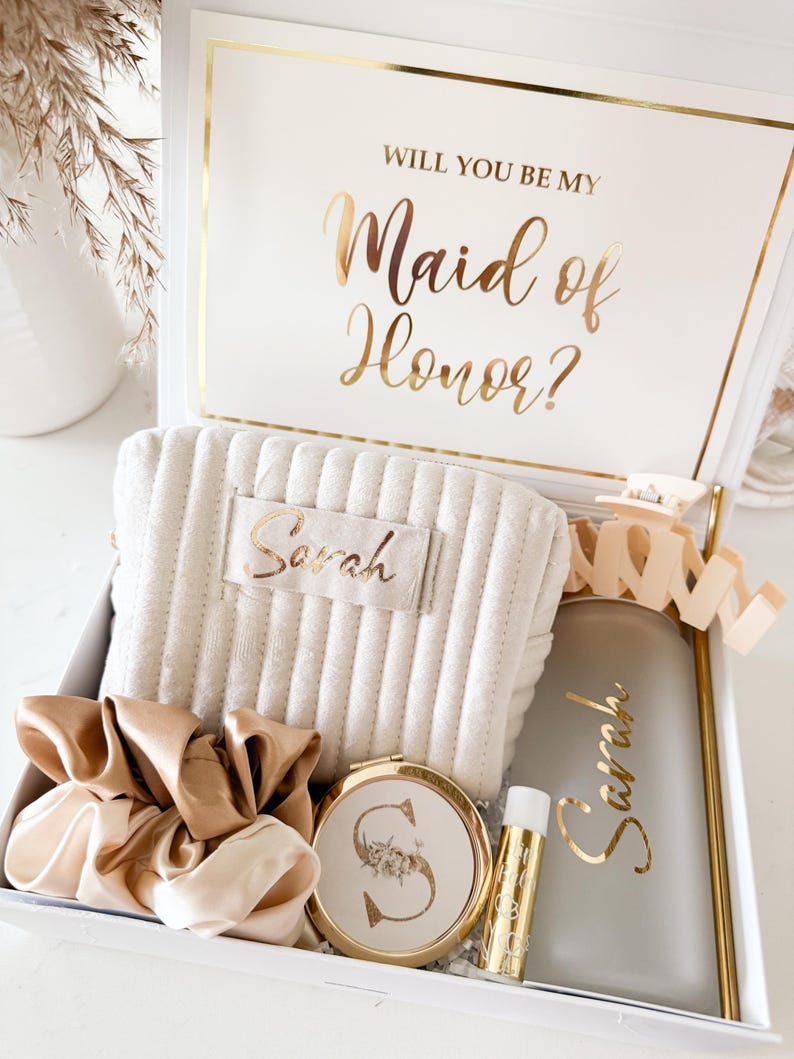 May include: A white gift box with gold trim, containing a beige quilted cosmetic bag, a gold compact mirror, a lip balm, a tumbler, and a scrunchie. The box lid reads "Will you be my Maid of Honor?" in gold script. The cosmetic bag and tumbler are personalized with the name "Sarah".