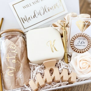 May include: A white gift box filled with items for a maid of honor. The box includes a glass tumbler with a gold straw, a white jewelry box with a gold initial "K", a gold pen, a gold hair tie, a white card with the text "I can't say I do without you", a white rose, and a wooden sign that says "Maid of Honor".