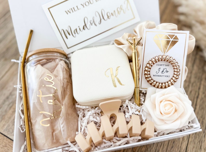 May include: A white gift box filled with items for a maid of honor. The box includes a glass tumbler with a gold straw, a white jewelry box with a gold initial "K", a gold pen, a gold hair tie, a white card with the text "I can't say I do without you", a white rose, and a wooden sign that says "Maid of Honor".