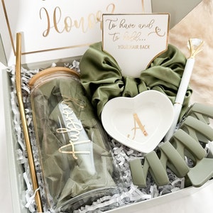 May include: A green and gold gift box filled with items for a bridesmaid. The box includes a tumbler with a gold lid and straw, a green scrunchie, a heart-shaped dish with a gold letter A, a green hair clip, and a pen with a diamond-shaped top.
