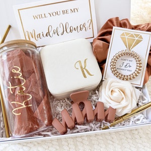 May include: A white gift box filled with items for a bridesmaid proposal. The box includes a personalized tumbler, a jewelry box, a hair scrunchie, a rose, a pen, and a card that says "Will you be my Maid of Honor?"