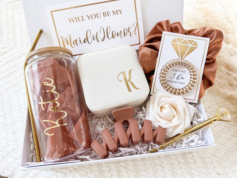 May include: A white gift box filled with items for a bridesmaid proposal. The box includes a personalized tumbler, a jewelry box, a hair scrunchie, a rose, a pen, and a card that says "Will you be my Maid of Honor?"