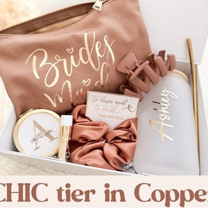 May include: A gift box with a copper theme, including a makeup bag with "Bridesmaid" in gold script, a hair clip, a tumbler with a gold straw, a compact mirror, lip balm, a scrunchie, and a card that says "To have and to hold... Your hair back."