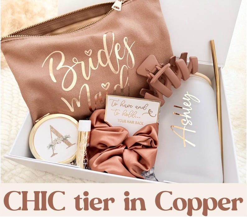 May include: A gift box with a copper theme, including a makeup bag with "Bridesmaid" in gold script, a hair clip, a tumbler with a gold straw, a compact mirror, lip balm, a scrunchie, and a card that says "To have and to hold... Your hair back."
